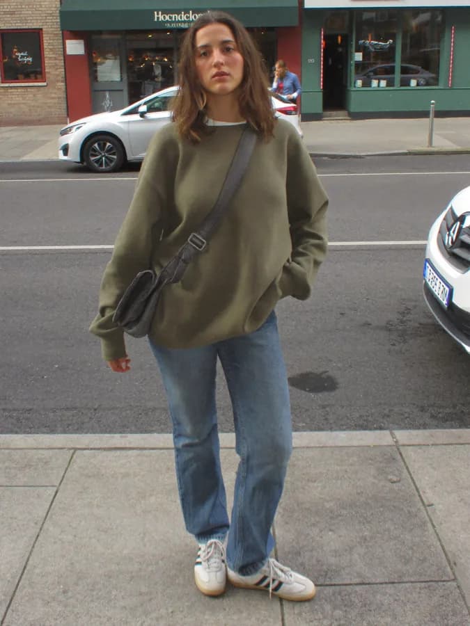 A woman on a pavement in an olive sweatshirt, jeans and white trainers
